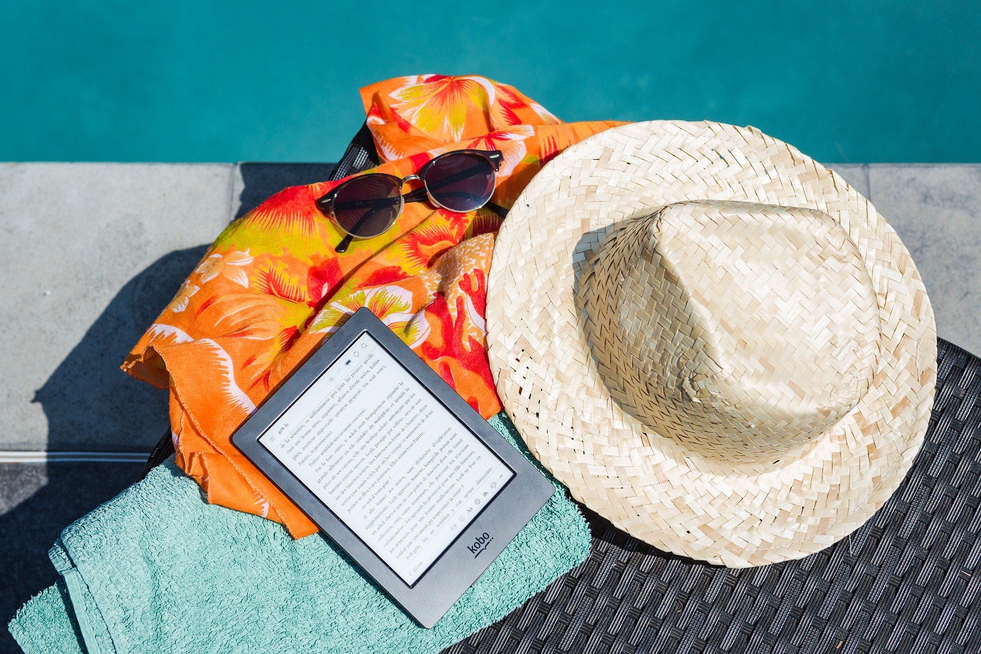 Kobo ereader, straw hat, orange swim cover up, blue towel, and sunglasses artfully laid by a pool's edge