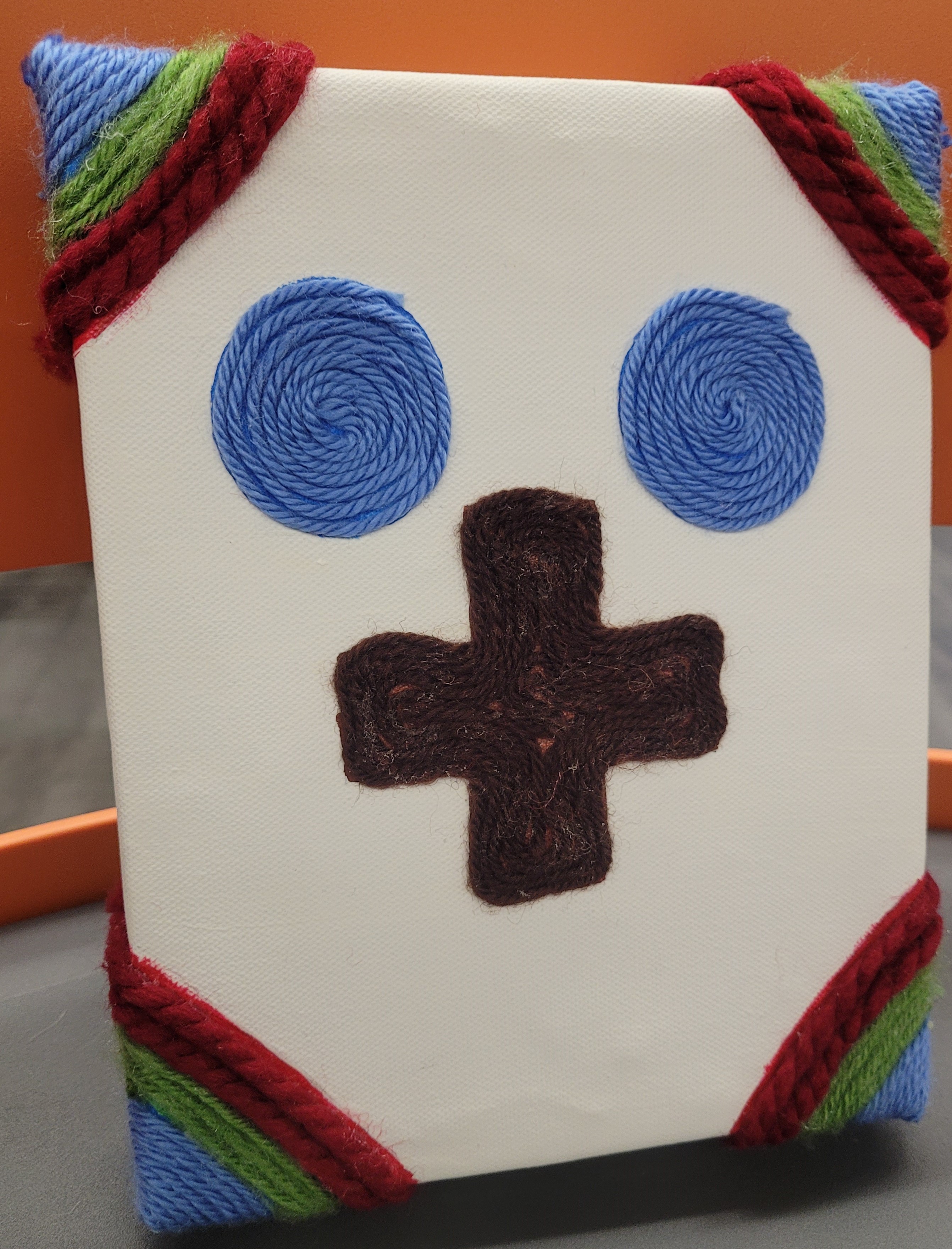 white canvas with green, red and blue yarn corners.  Two blue yarn circles on the top center and a brown yarn plus sign in the middle.
