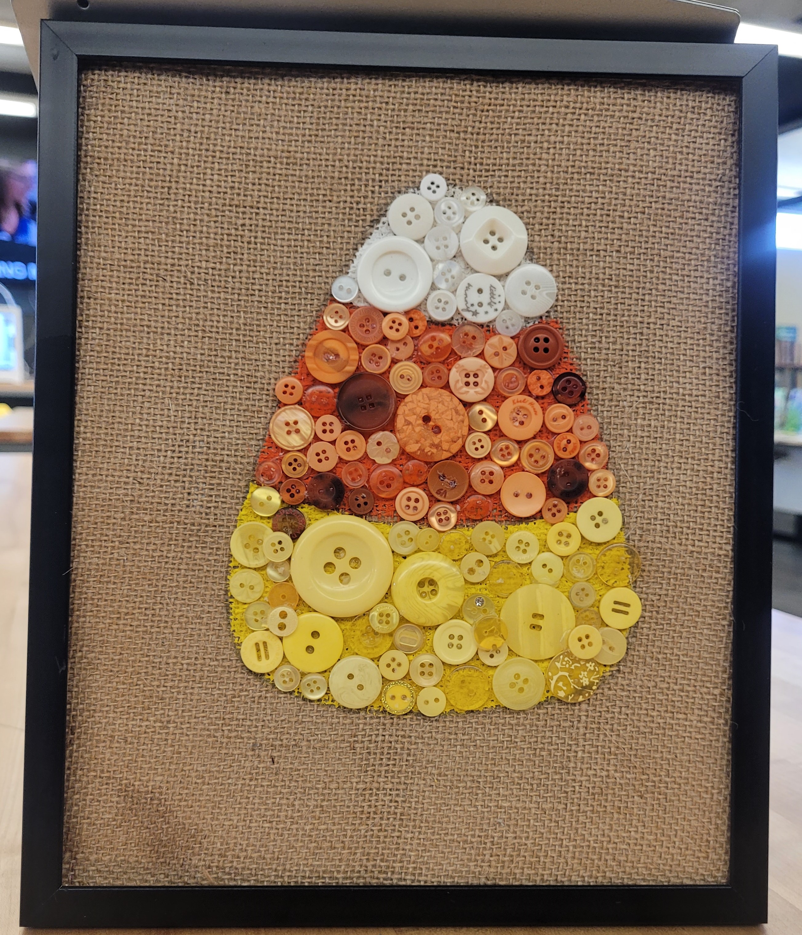 white, orange, and yellow candy corn filled in with white, orange and yellow buttons on a burlap back in a black picture frame.