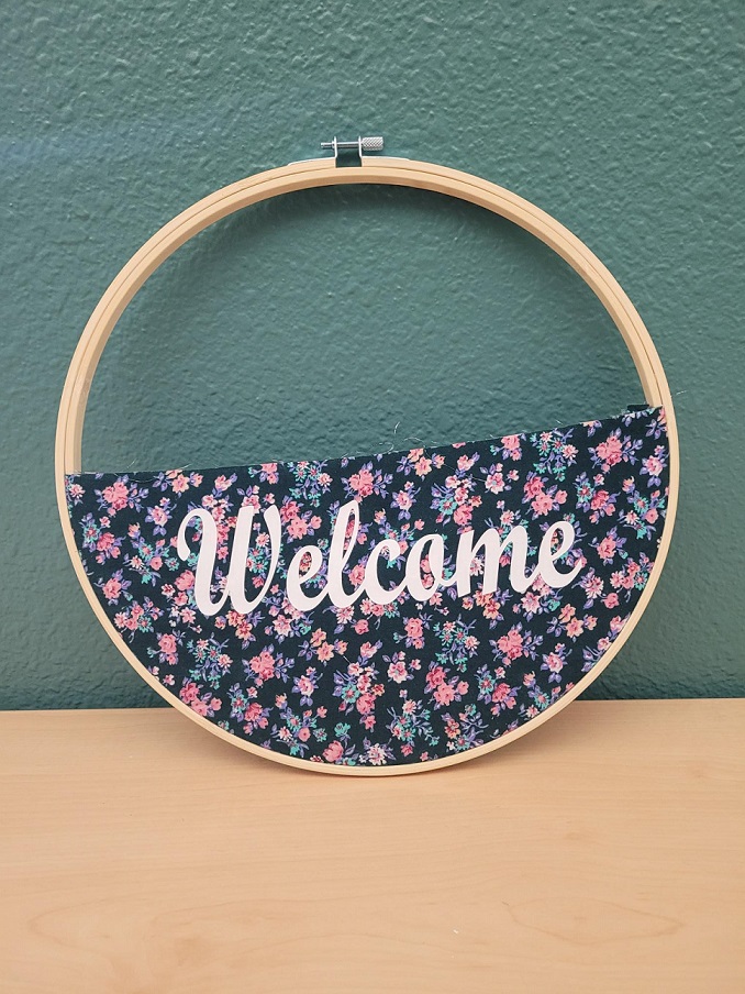Wooden embroidery hoop with a floral fabric pocket halfway up the inner circle. In the middle of the pocket is a white vinyl sticker that says "Welcome."