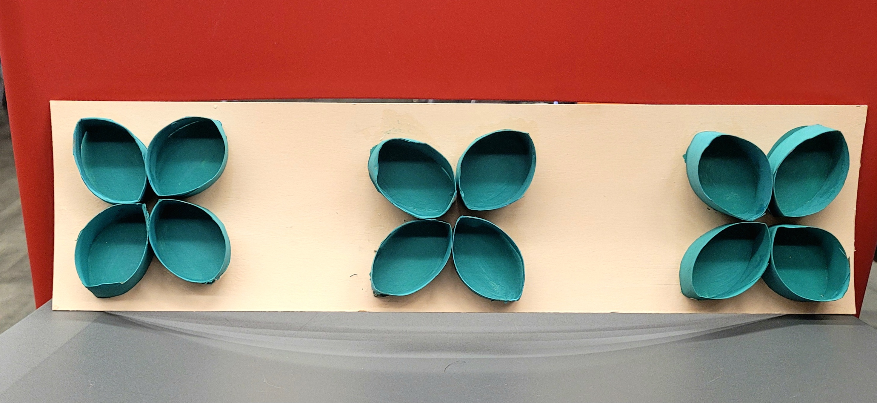 blue/green painted cardboard roll pieces in a flower pattern on a cream, rectangle backing.