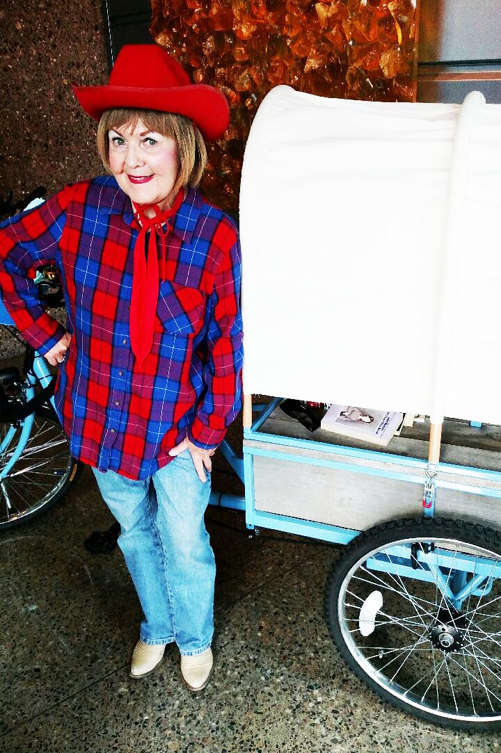 Woman dressed as cowgirl standing next to a wagon hitched to a bicycle.