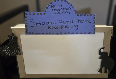 Cardboard box with a blue sign that states red mountain shadow puppet theater now playing with a dancing couple silohouette and an elephant silhoutte.