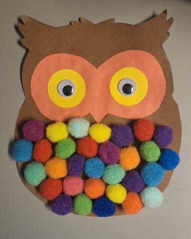 A brown paper owl with multicolored pom-poms and orange and yellow eyes.