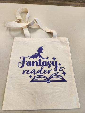 A tote bag with a dragon and book design which reads Fantasy Reader. The design is purple and the bag is a beige color.