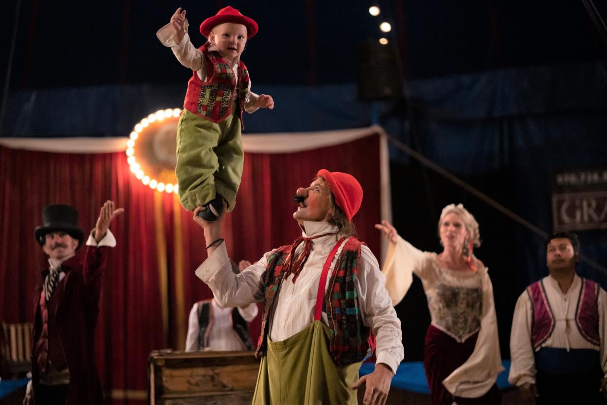 Circus scene with a male clown holding a toddler clown on his fingertips.