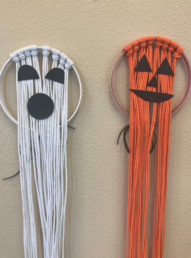 There is a white circle with white macrame cord knotted around the top of the hoop with the threads draping down. There are two black eyes and a black circle for a mouth to resemble a ghost. The second object is similar except the circle and macrame cord is orange and there is a small triangle nose to resemble a pumpkin.